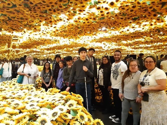 A group of teenagers and adults stand surrounded by yellow sunflowers looking into the camera. The teenage boy in the middle is holding a cane.