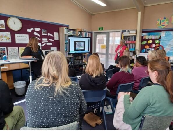 A group of people looking towards a large TV screen where information is being presented by two presenters standing on either side