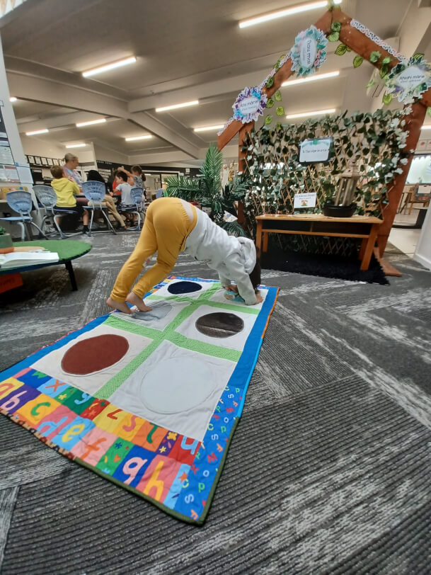 Child is on all fours on the Twister game mat on the floor. She is making an ‘i’ with her body. Her legs are on Dot 2, and her arms in Dot 4. Each dot is made from a different texture fabric.
