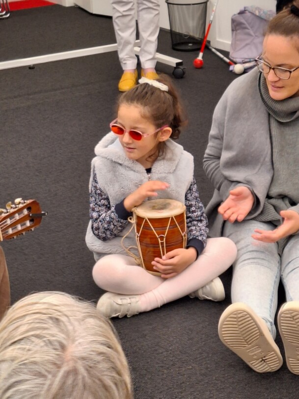 Dima is sitting on the floor playing a hand drum alongside a first year Music therapist.