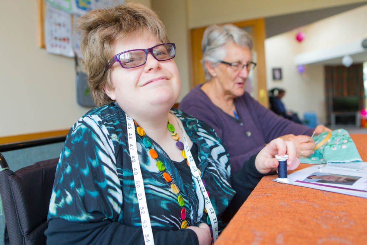 Katelyn sits at a table with her carer who is stitching.