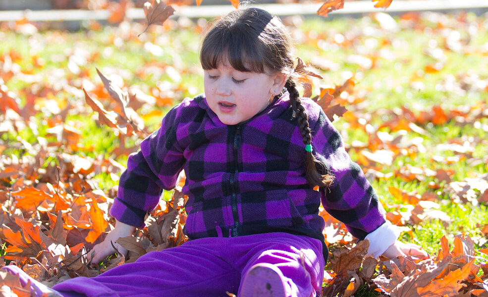 Giana sitting outside in the sun surrounded by autumn leaves.