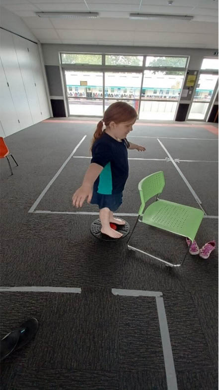Laura is in a classroom standing on a balance board practising balancing activities with her RTV prior to the Athletics Day.