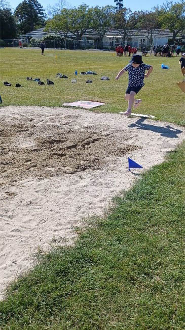Laura is making a jump on the long jump track on the grass and sand.