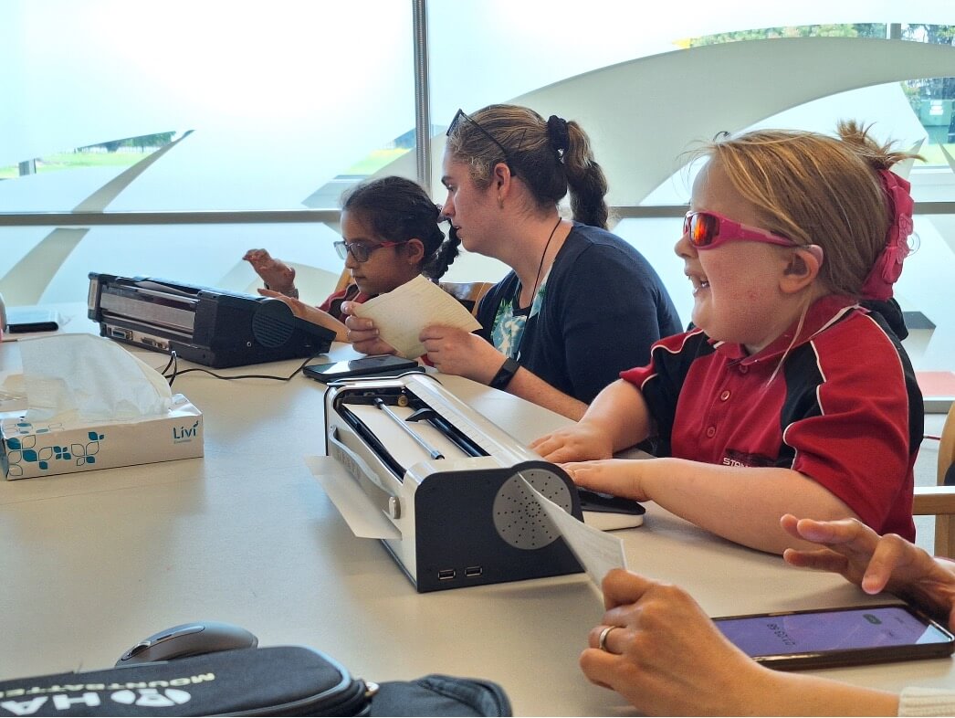 One female Resource Teacher Vision sitting between two young girls who are typing on their braille device.