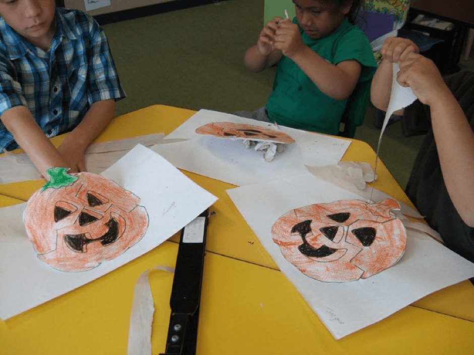 Three children tearing strips of paper to stuff into pumpkin shapes.