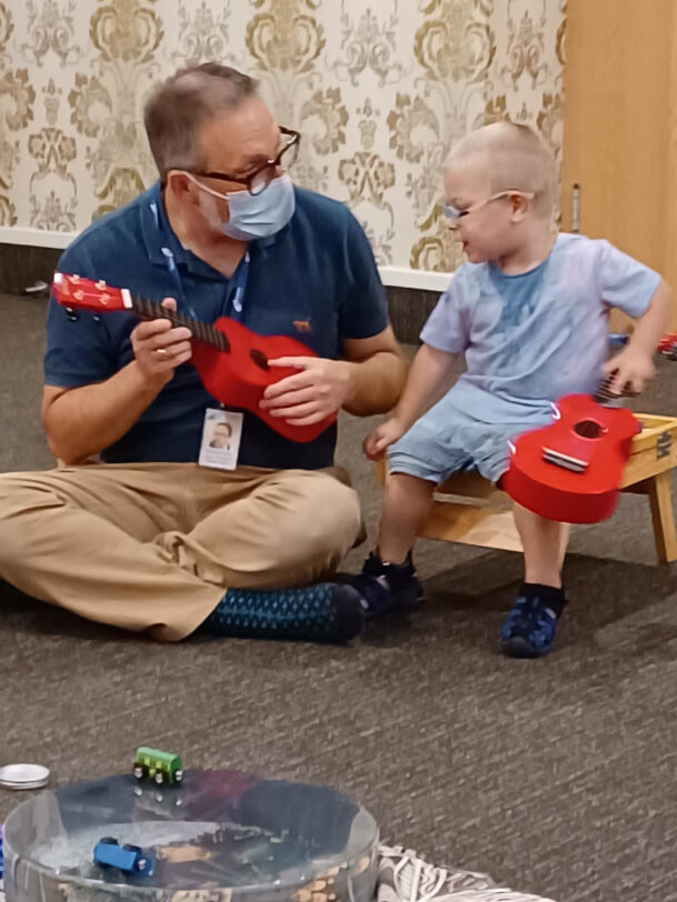 Patrick sitting on the floor with Hunter exploring the ukulele.