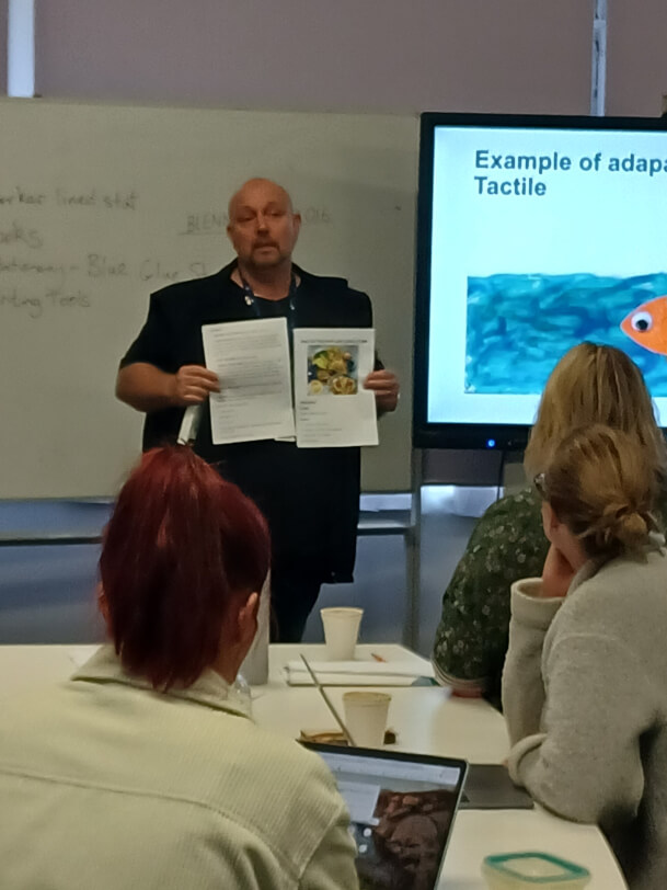 TV is standing talking to a group of attendees at a workshop about adapting resources for visually impaired learners.