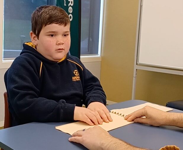 Reid is sitting in a chair at a table exploring shapes in a book with thermoform pages.