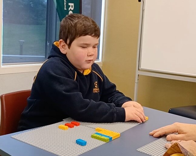 Reid is sitting in a chair at a table using LEGO Braiile Bricks to write his name on a board.