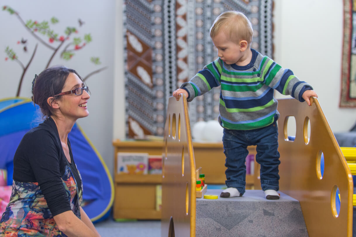 Robert stands in the middle of climbing bench looking to his right at mum who sitting beside.