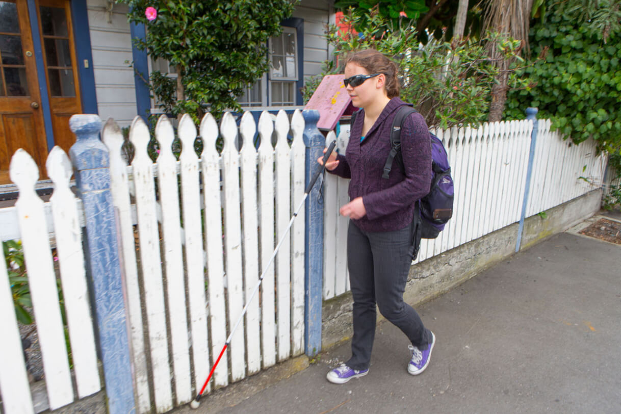 Mary walking in front of a house using her cane.