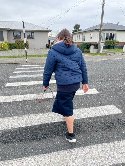 Tahlia is crossing the road on a zebra crossing using her cane.
