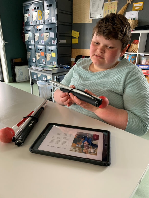 rinity is sitting at a table holding a cane in her hands and looking at a ipad during a zoom session.