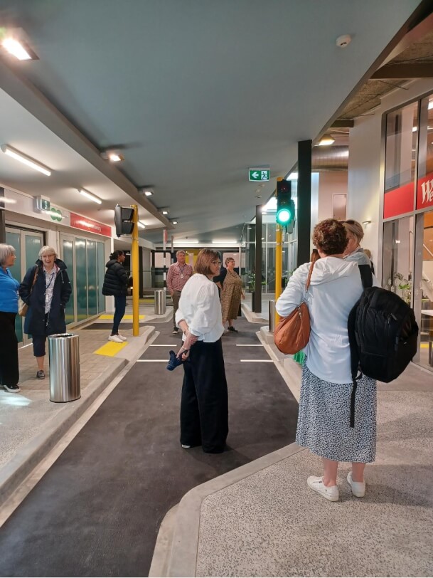 Nine adults talking to each other, while exploring the traffic lights, escalator, lifts, bus and train at the mobility centre at BLVNZ.
